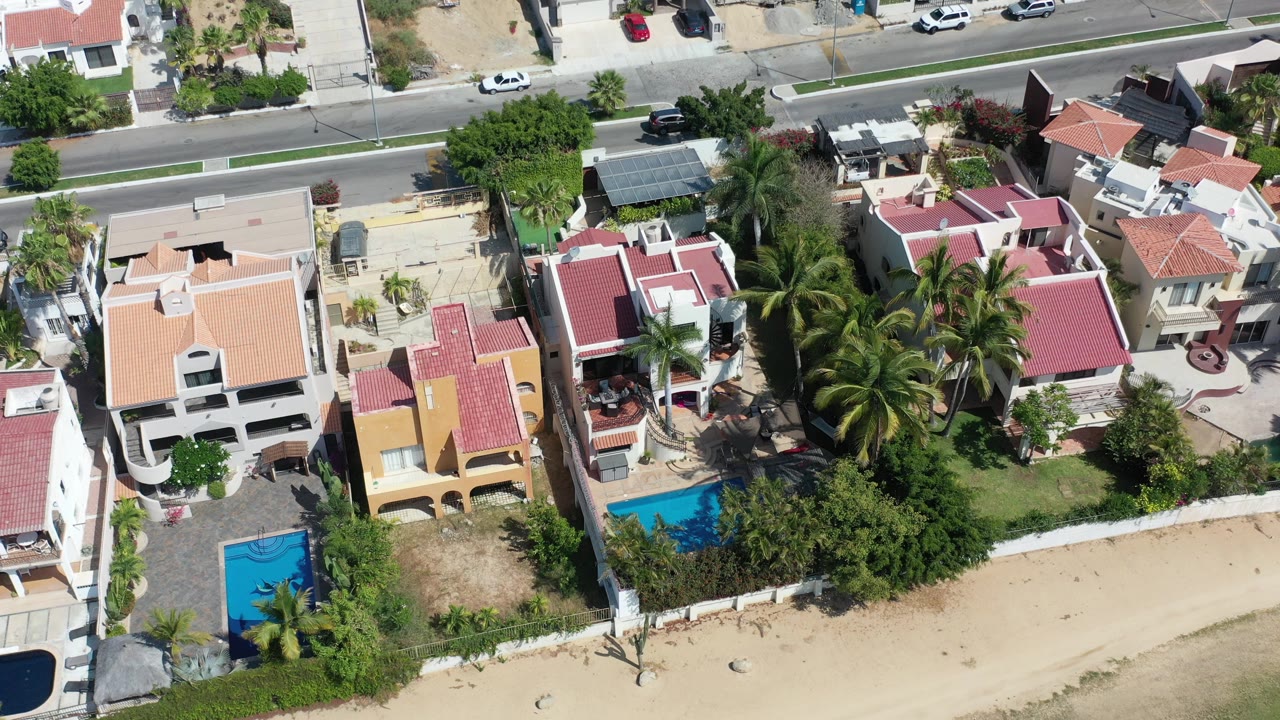Opening overhead frame of Los Cabos homes with red and tan roofs, two blue pools, palm trees, sandy ground, a paved road, and no visible faces.