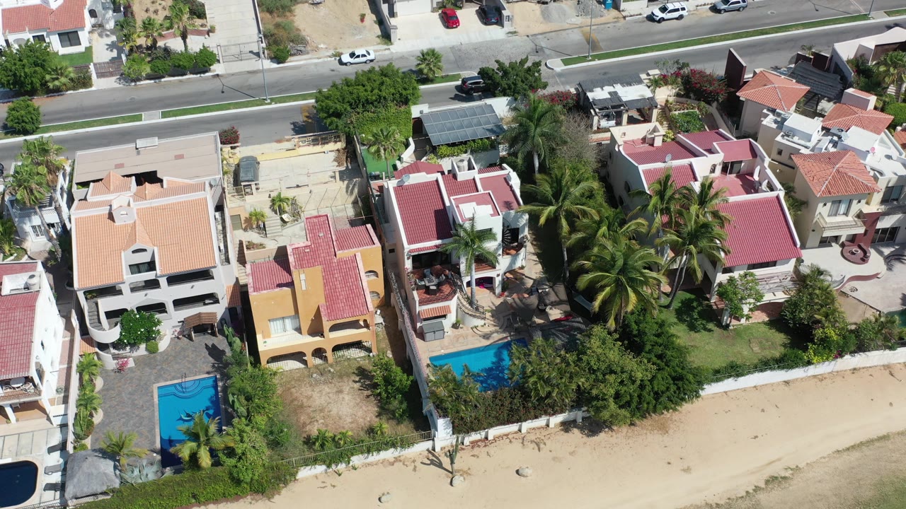 Los Cabos aerial frame showing neighboring homes, a bright blue pool at lower left, palms between yards, sandy open ground, and parked cars along the road.