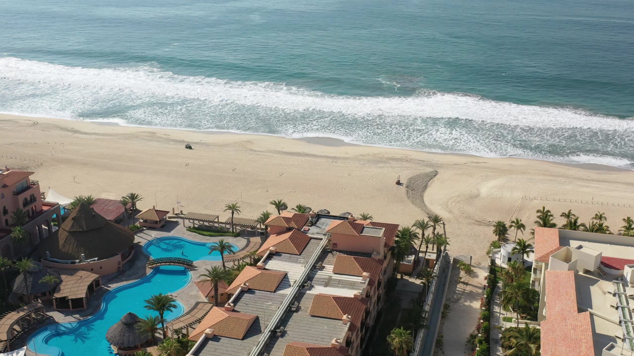 Opening aerial frame over Los Cabos, Mexico, with blue pools and red-tile villa rooftops in the foreground, pale sand and breaking surf beyond, and tiny distant people on the beach.