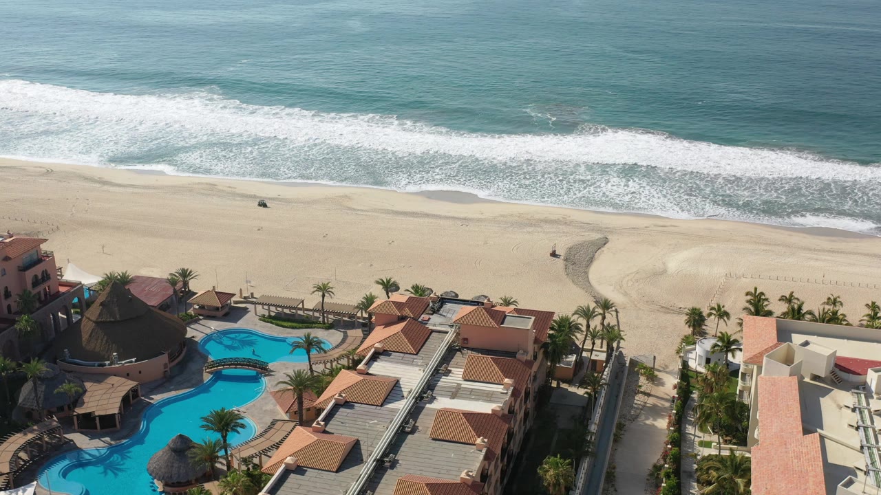 Los Cabos aerial view with foamy waves crossing the upper frame, a broad sand beach in the middle, and turquoise pools, palms, and villa roofs along the lower edge.
