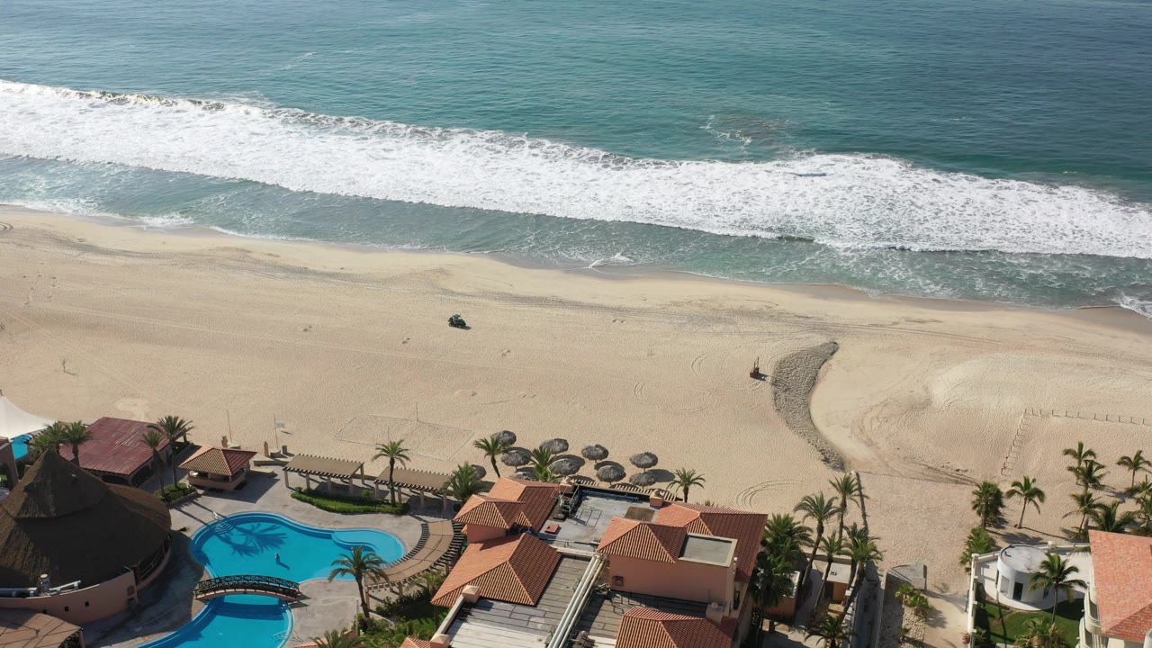 Aerial beach view with surf, homes, pool, palms, umbrellas, and a dark vehicle on sand.
