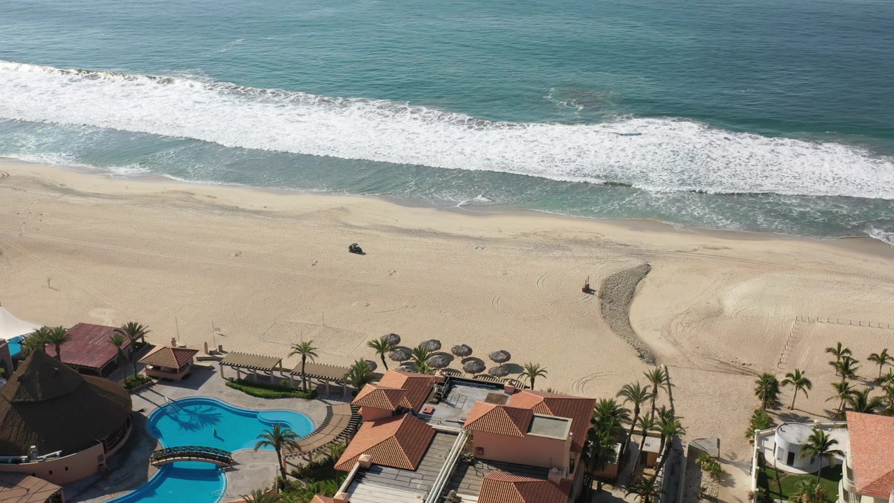 A slightly later aerial frame in Los Cabos, Mexico, with the dark vehicle still on the sand, white surf rolling across the top of frame, beachfront roofs and pool at the bottom, and no visible people.