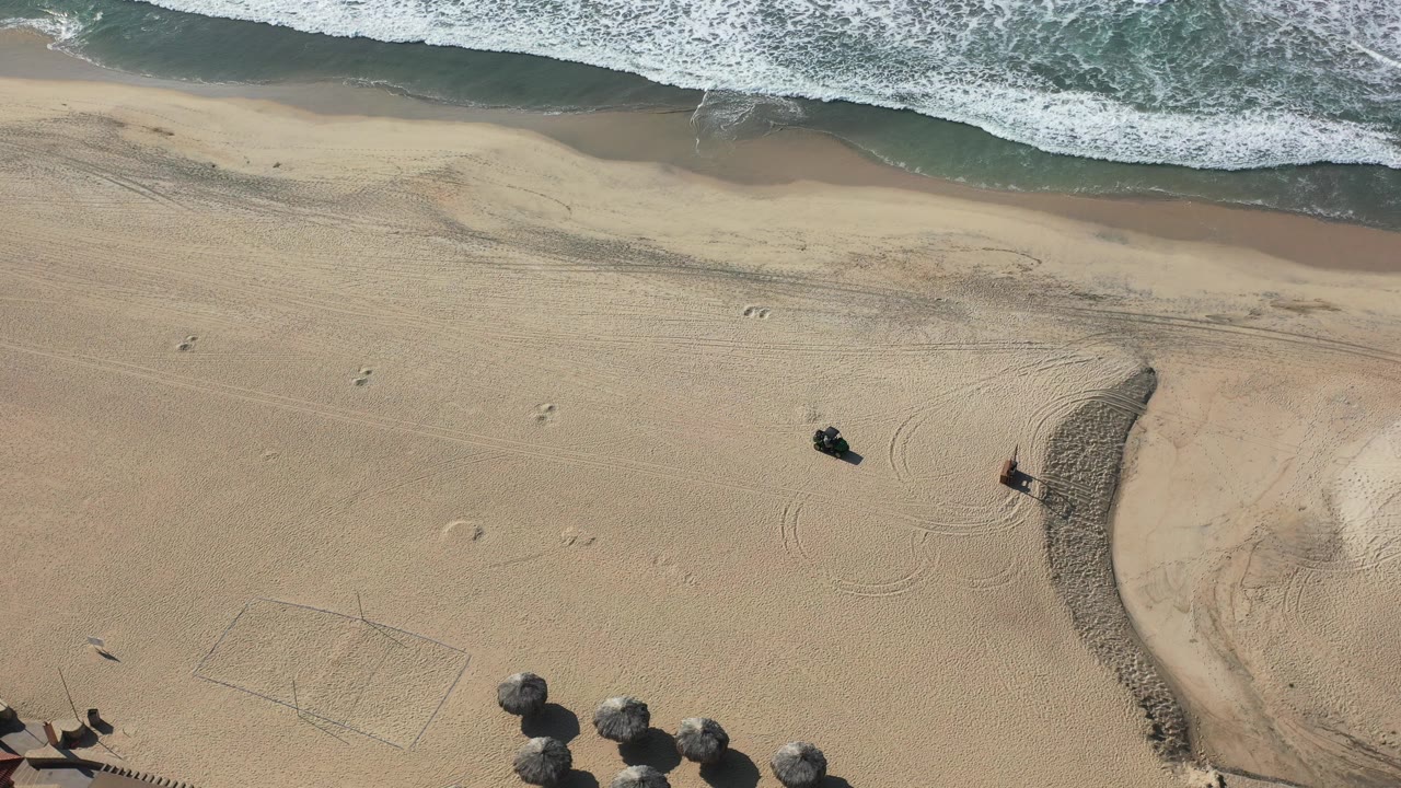 Overhead beach with surf, sand tracks, and small figures near a curved sand berm.