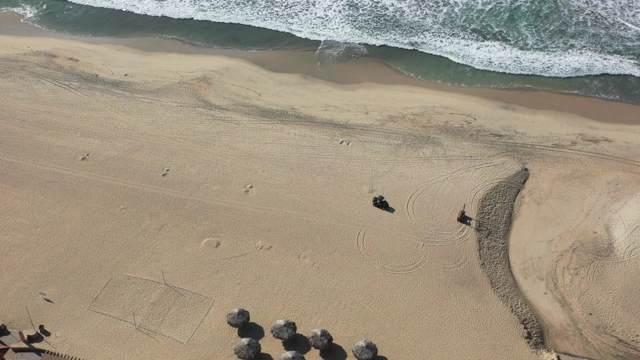 Overhead Los Cabos beach frame at 0:01, with the surf band across the top, textured sand through the center, footprints, and small people visible near the lower beach area.