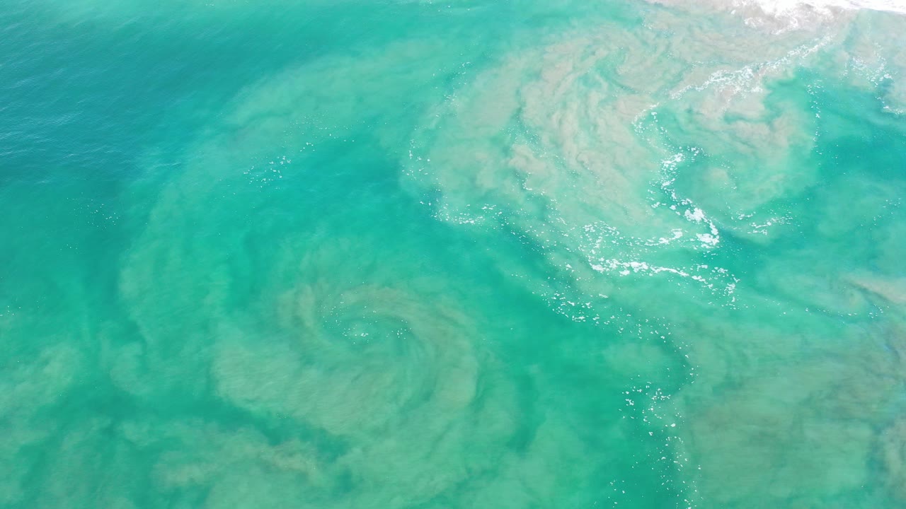 Overhead view of turquoise ocean water in Los Cabos, Mexico, with pale suspended sand patterns below the surface and no visible people.