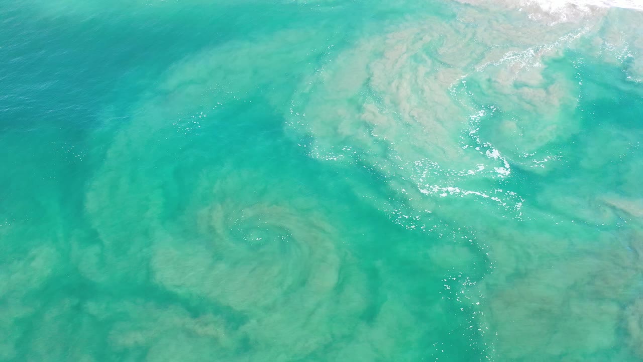 Aerial frame over Los Cabos water with green-blue tones, cloudy sand movement below, small foam flecks, and no visible people.