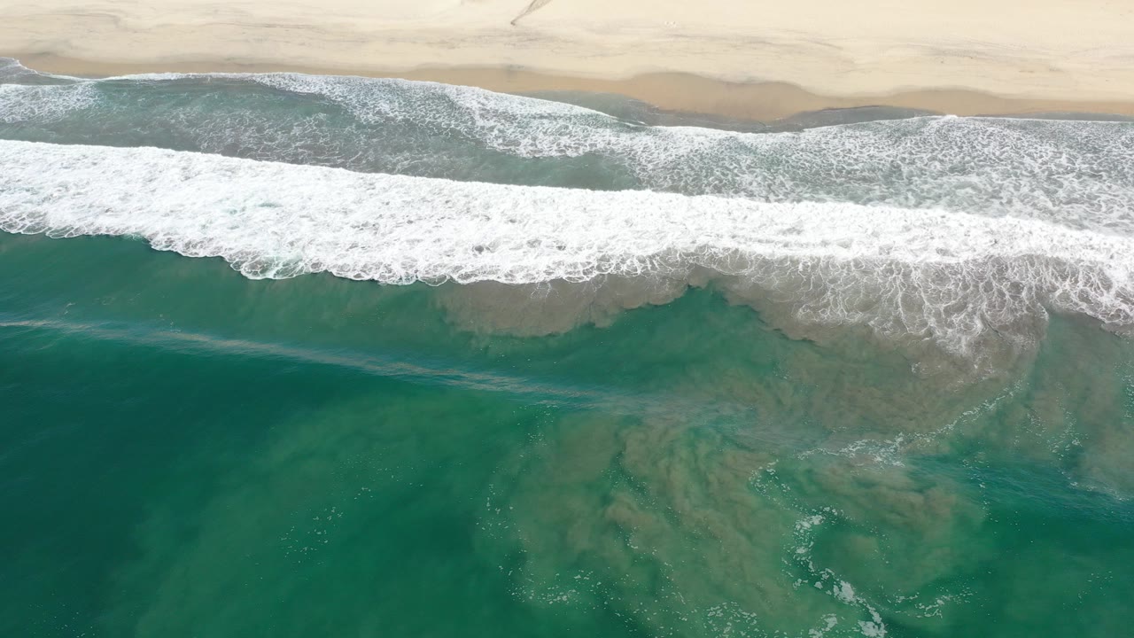 Overhead view in Los Cabos, Mexico, with teal-green ocean and white surf rolling toward a wide sand beach, no visible people.