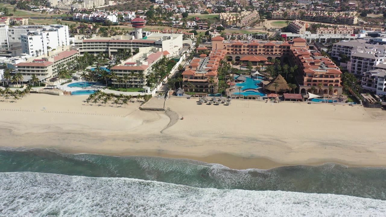 High aerial frame over Los Cabos, Mexico, with white surf crossing the lower edge, a broad sandy beach, resort buildings, blue pools, and inland development beyond, with no visible people.