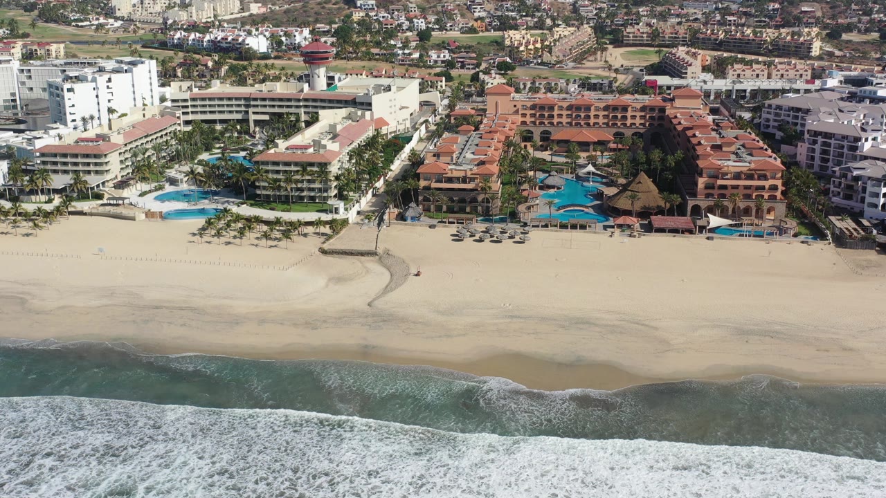Aerial view in Los Cabos, Mexico, showing foamy shoreline water at bottom, pale sand across the center, white hotel blocks, red-roofed buildings, pools, palms, and no visible people.