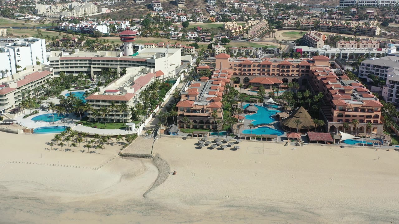 Aerial frame over Los Cabos beachfront hotels with red tile roofs, blue pools, palm-lined grounds, wide sand in front, dense buildings behind, and no visible people.