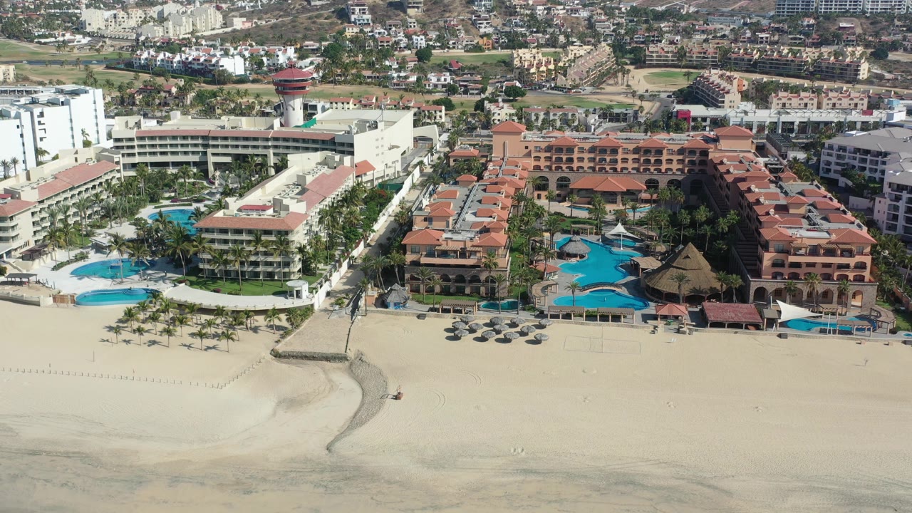 Aerial view in Los Cabos showing the sandy shoreline lower in frame, a white hotel building on the left, red-roofed lodging on the right, blue pools, palms, and no visible people.