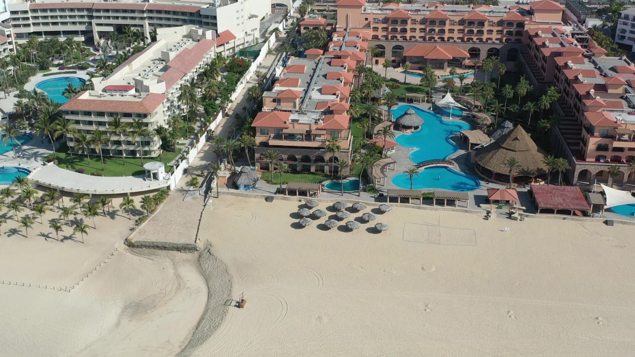 Aerial view in Los Cabos, Mexico, with the hotel pool area centered, palapa-style shade roofs near the water, beach sand in front, and no visible people.