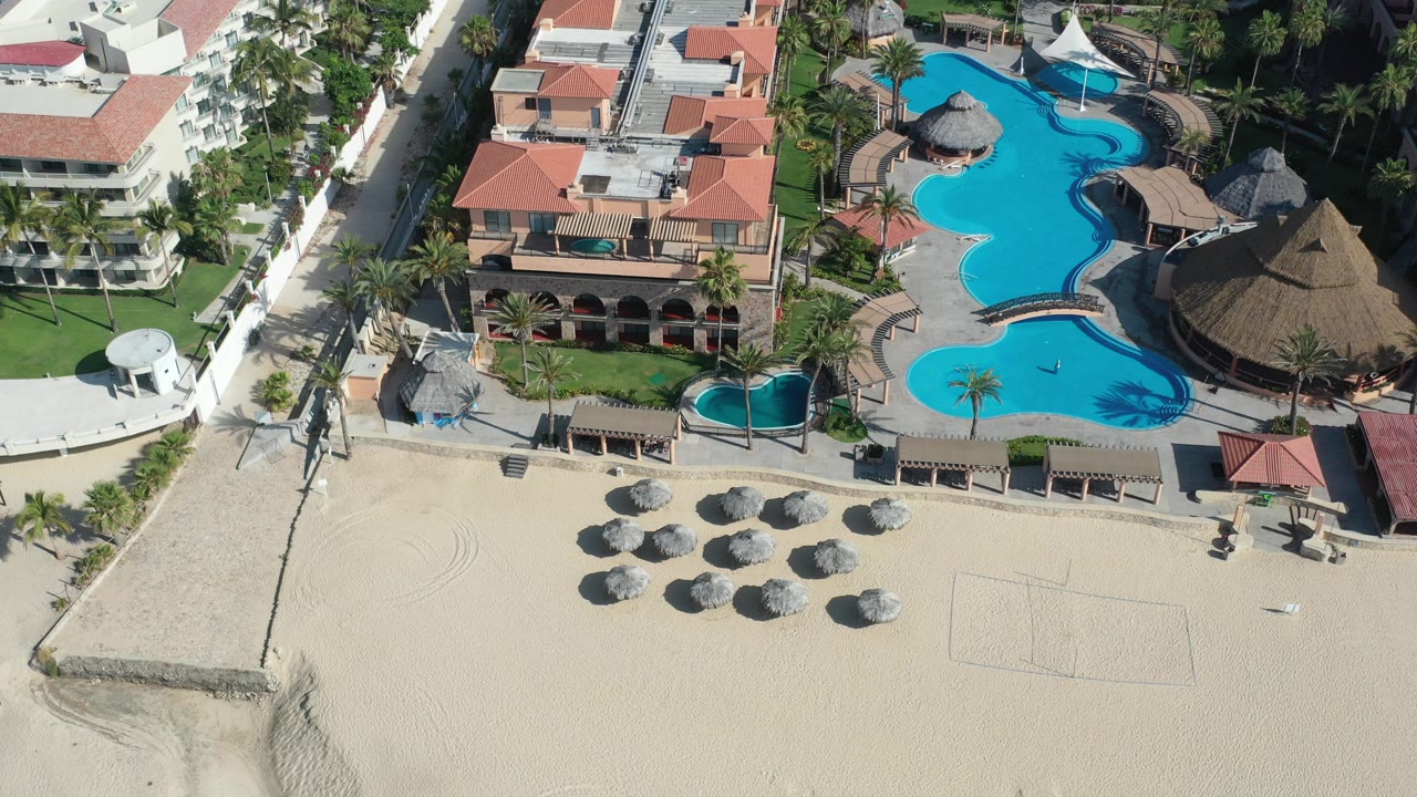 High-angle aerial frame over a Los Cabos beachfront resort, with turquoise lagoon pools, red tile roofs, palapa umbrellas on sand, palms, and no visible people.