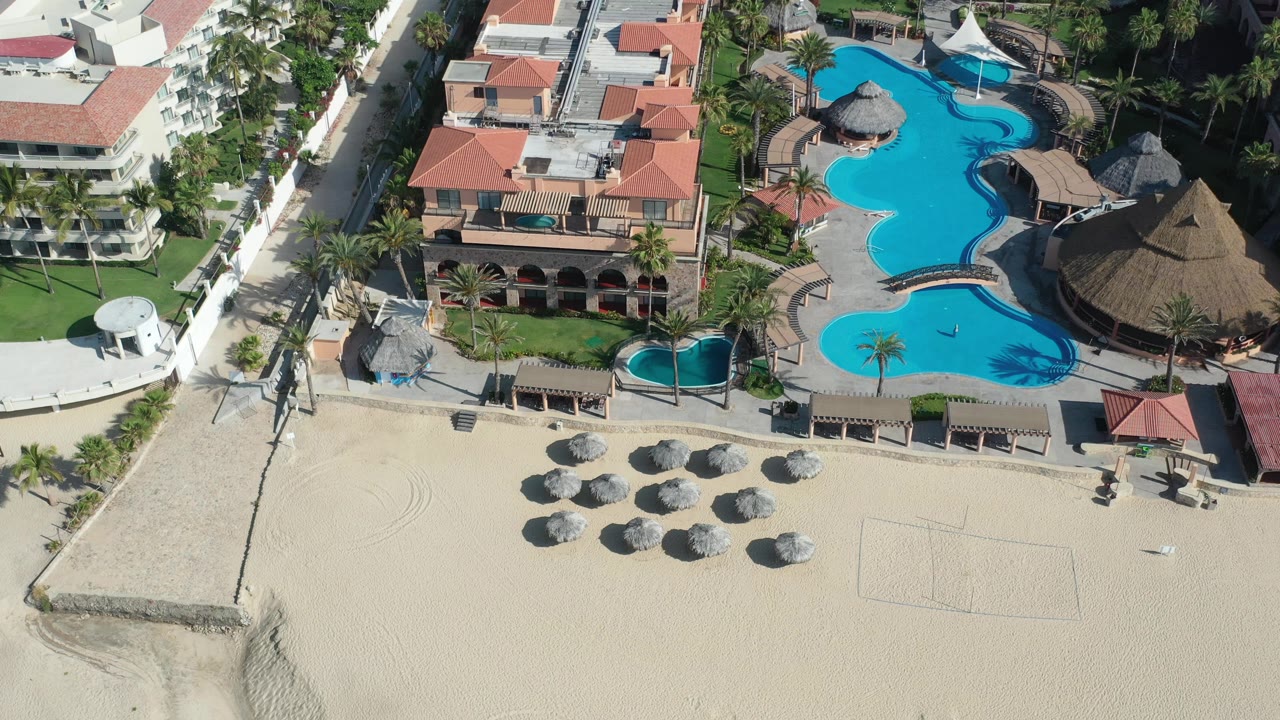 Aerial view in Los Cabos showing the resort buildings centered above a sandy beach, with curved blue pools to the right and no visible people.