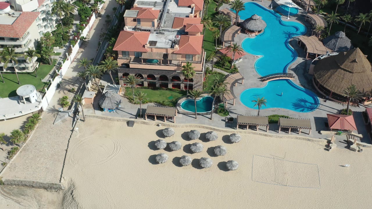 Drone view of resort buildings, palms, sand, and lagoon-style pools.