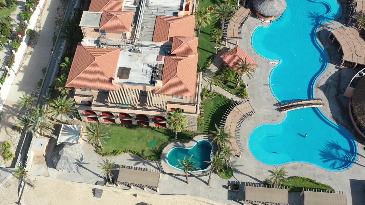Overhead view of a Los Cabos resort with terracotta roofs, bright blue pools, palm trees, lawn, cabanas, and no visible people at the opening moment.