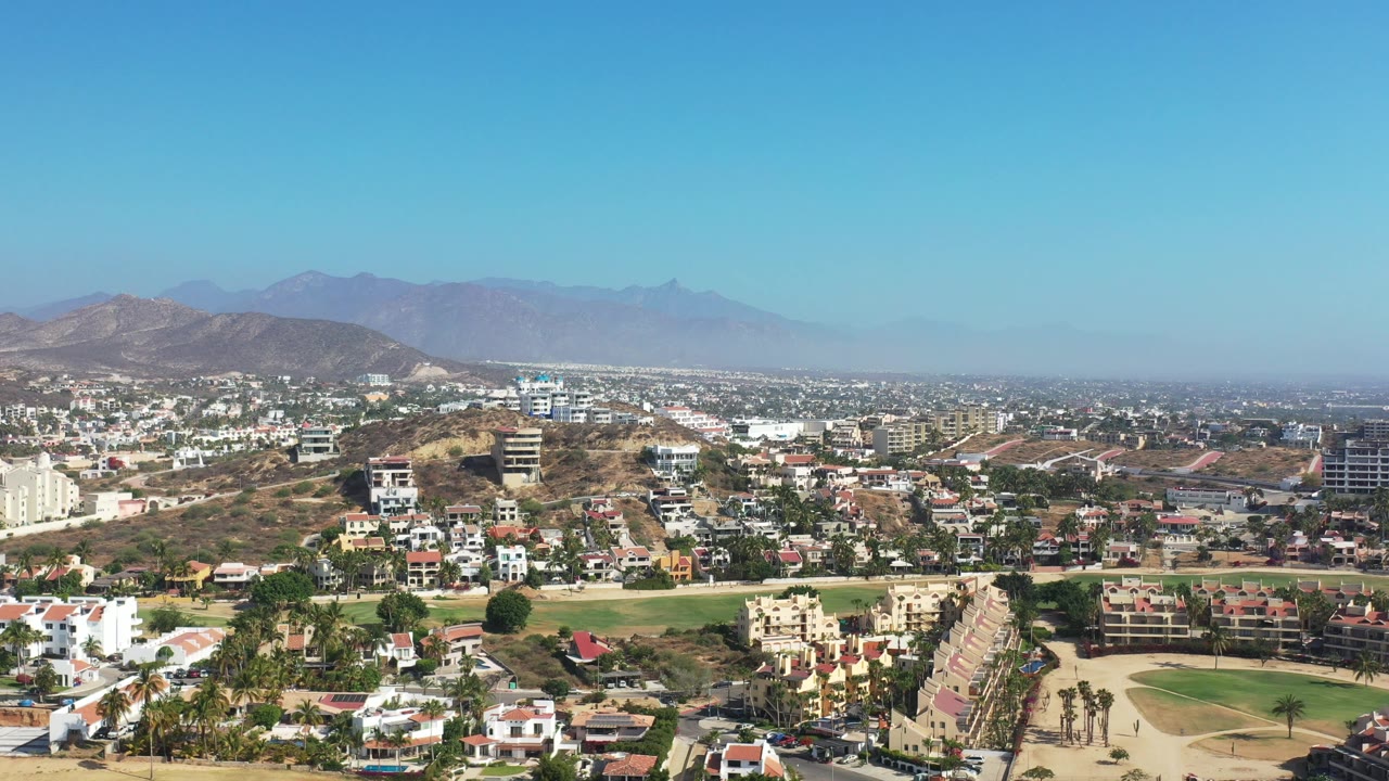 Opening aerial frame over Los Cabos, Mexico, with homes, roads, a green field, dry hillside terrain, distant mountains, blue sky, and no visible people.