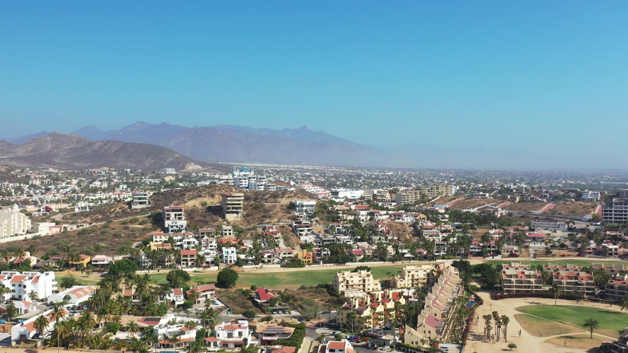Wide aerial of neighborhoods, green space, and mountains under blue sky.
