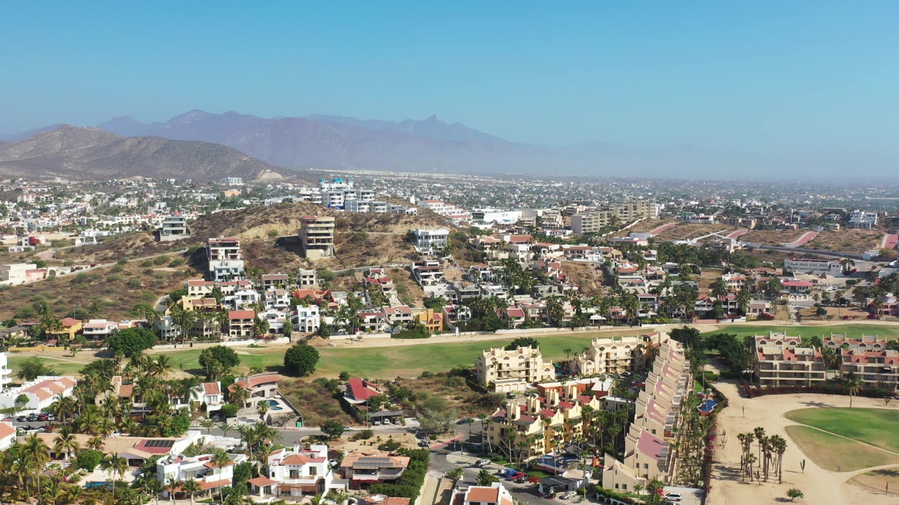 High view across Los Cabos residences and palms, with tan hills in the foreground, a broad city spread in the distance, and no visible people.