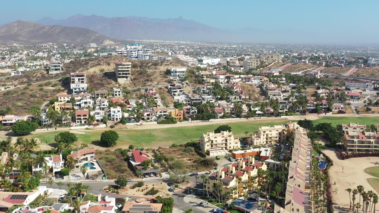 Opening aerial frame over Los Cabos, Mexico, showing hillside residences, a green strip, palms, dry slopes, distant mountains, hazy sky, and no visible people.