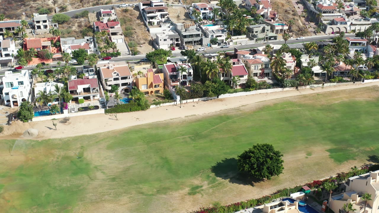 Los Cabos aerial frame showing clustered villas, a road with vehicles, a sandy border, green open land, and a large tree near the lower right, with no visible people.