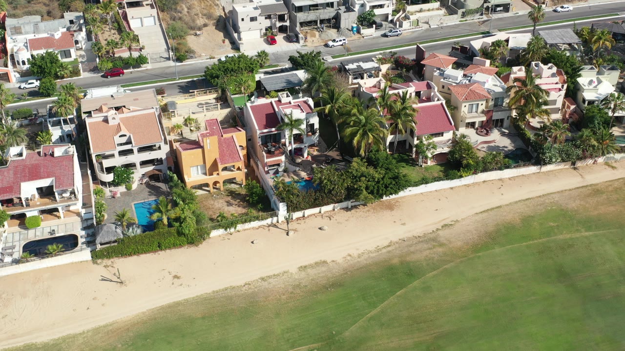 High-angle aerial frame over Los Cabos beachfront homes with private pools, palm trees, a sandy strip, green nearshore water, and no visible people.