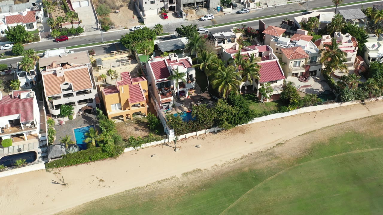 Coastal homes, palm trees, pools, beach, and road from above.