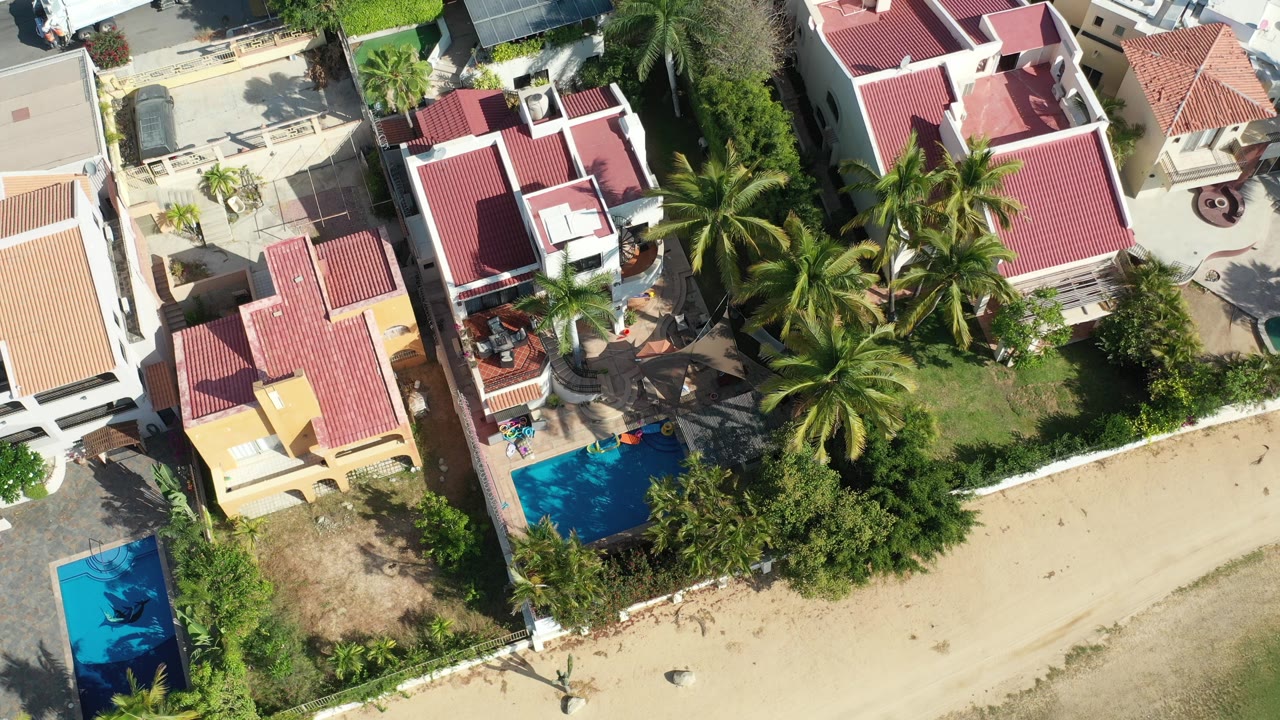 Overhead view of red-roofed homes beside pools, palms, and a sandy beach edge.
