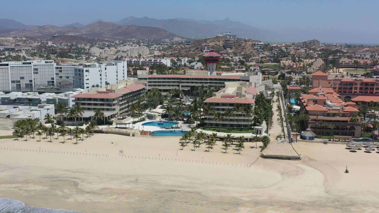 Opening aerial frame over Los Cabos, Mexico, showing pale beach foreground, hotel buildings with pools and palms, inland town blocks, distant dry hills, and no visible people.