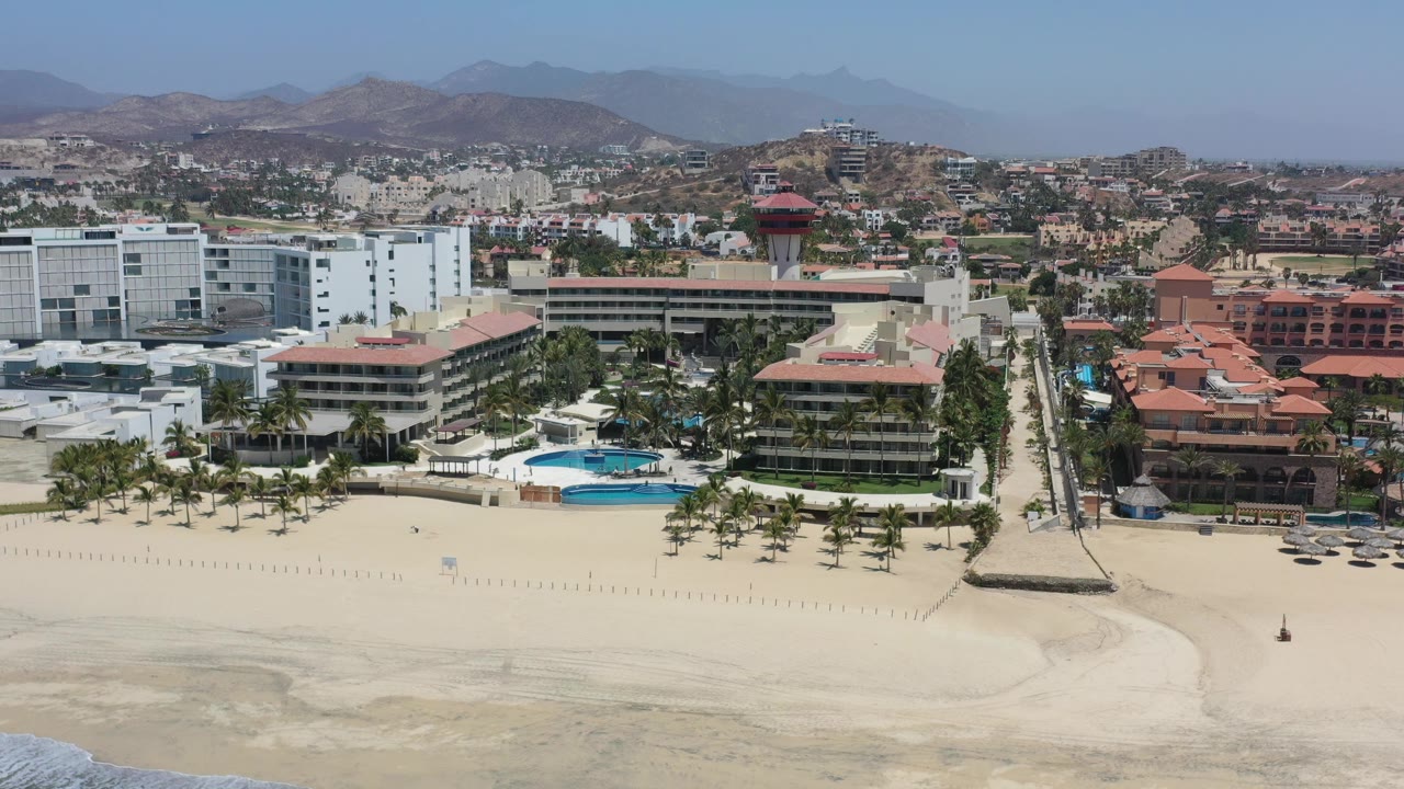 Aerial frame in Los Cabos, Mexico, with the beach across the lower frame, white and red-roofed buildings beside pools, a central walkway, inland neighborhoods, hills, and no visible people.