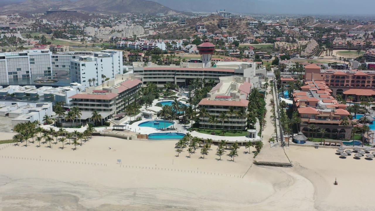 Aerial view of beachfront resorts, sand, pools, palms, town, and hills.