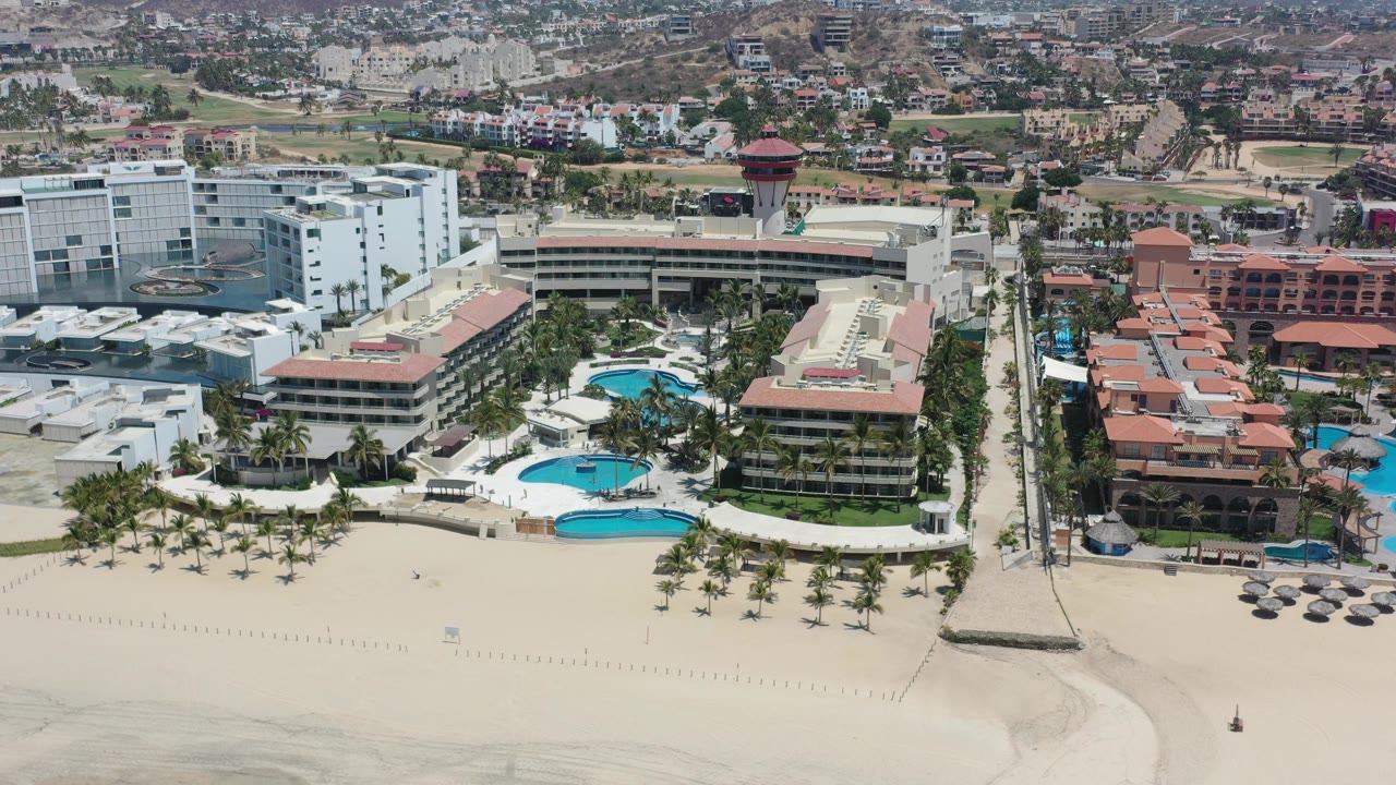 Aerial view of beachfront hotels, pools, palms, and pale sand.