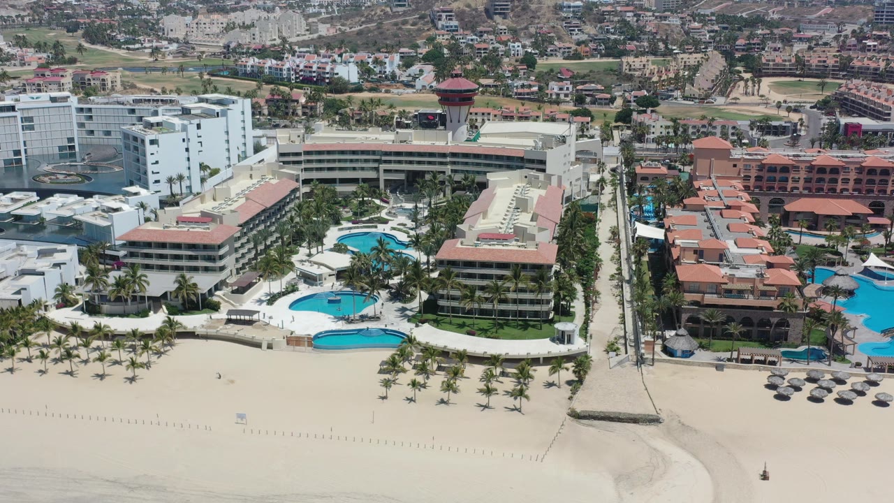 Opening aerial frame of a Los Cabos beachfront hotel area with white buildings, blue pools, palm-lined sand, and no visible people.
