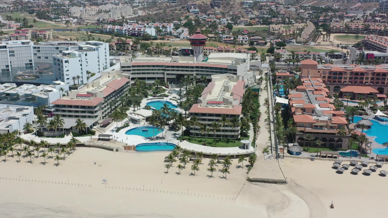 Aerial frame in Los Cabos showing curved blue pools beside white hotel blocks, red roof structures, beach sand, and no visible people.