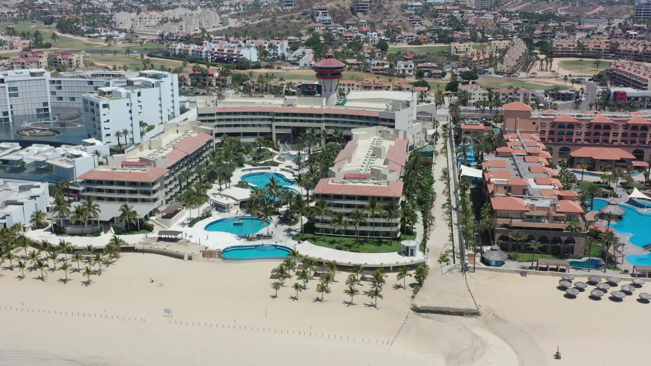 Drone-height view over hotel roofs, pool courtyards, beach, and palms.