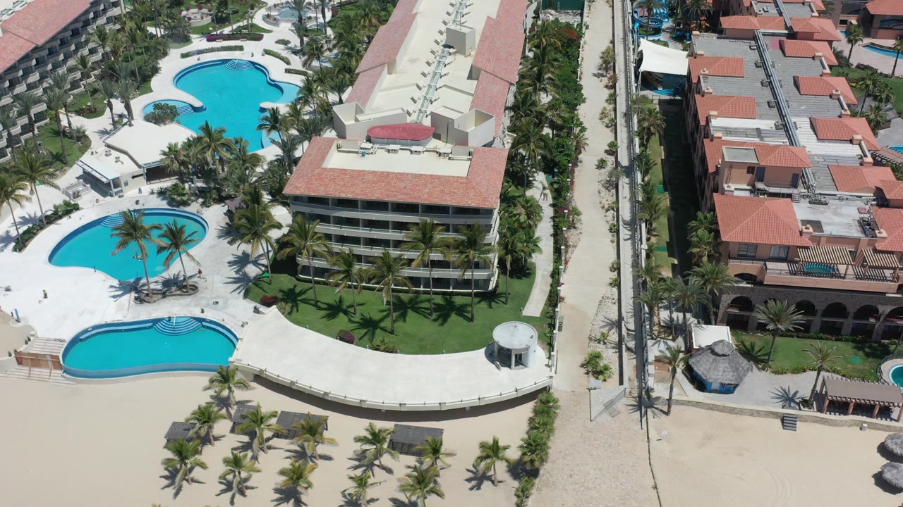 Aerial frame in Los Cabos, Mexico looking over curved blue pools, palm landscaping, red roof structures, a narrow street, and beachfront sand with no visible people.
