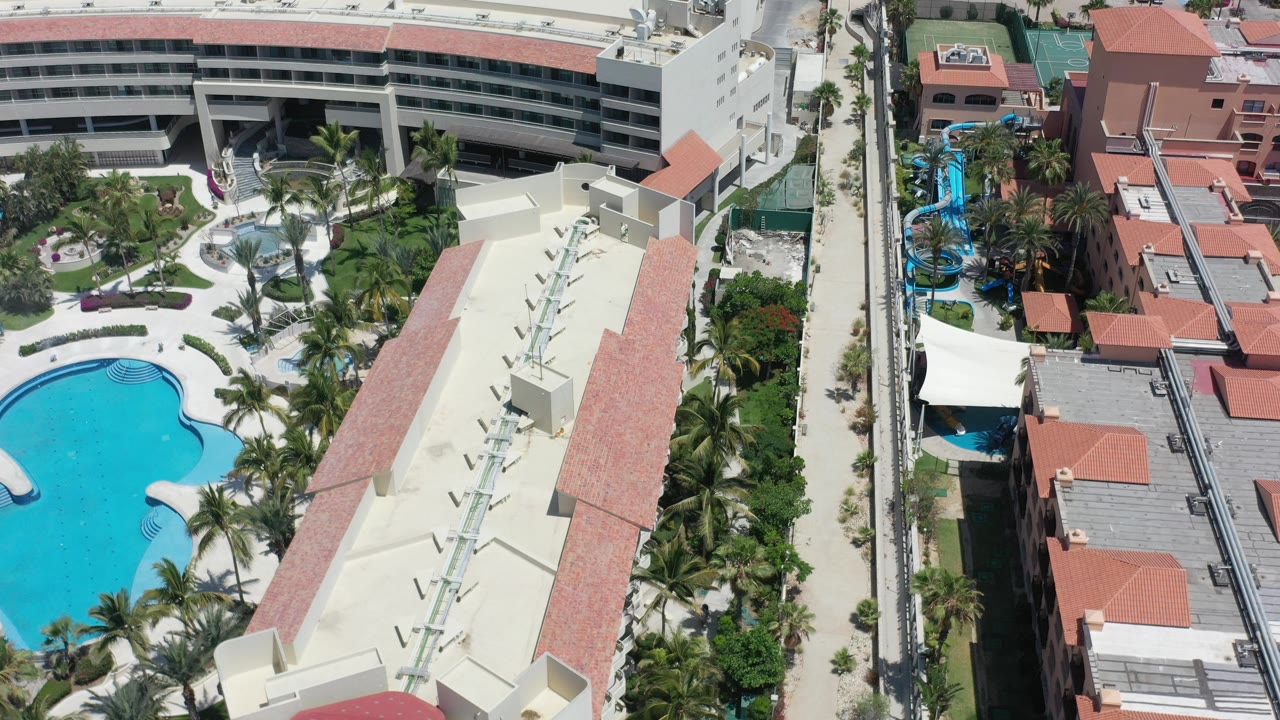 Aerial view over a Los Cabos resort pool, cream buildings, terracotta roofs, palms, and walkways in bright daylight, with no visible people.