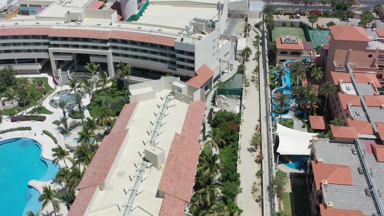 Aerial view of curved resort building, blue pool, palms, and neighboring rooftops.