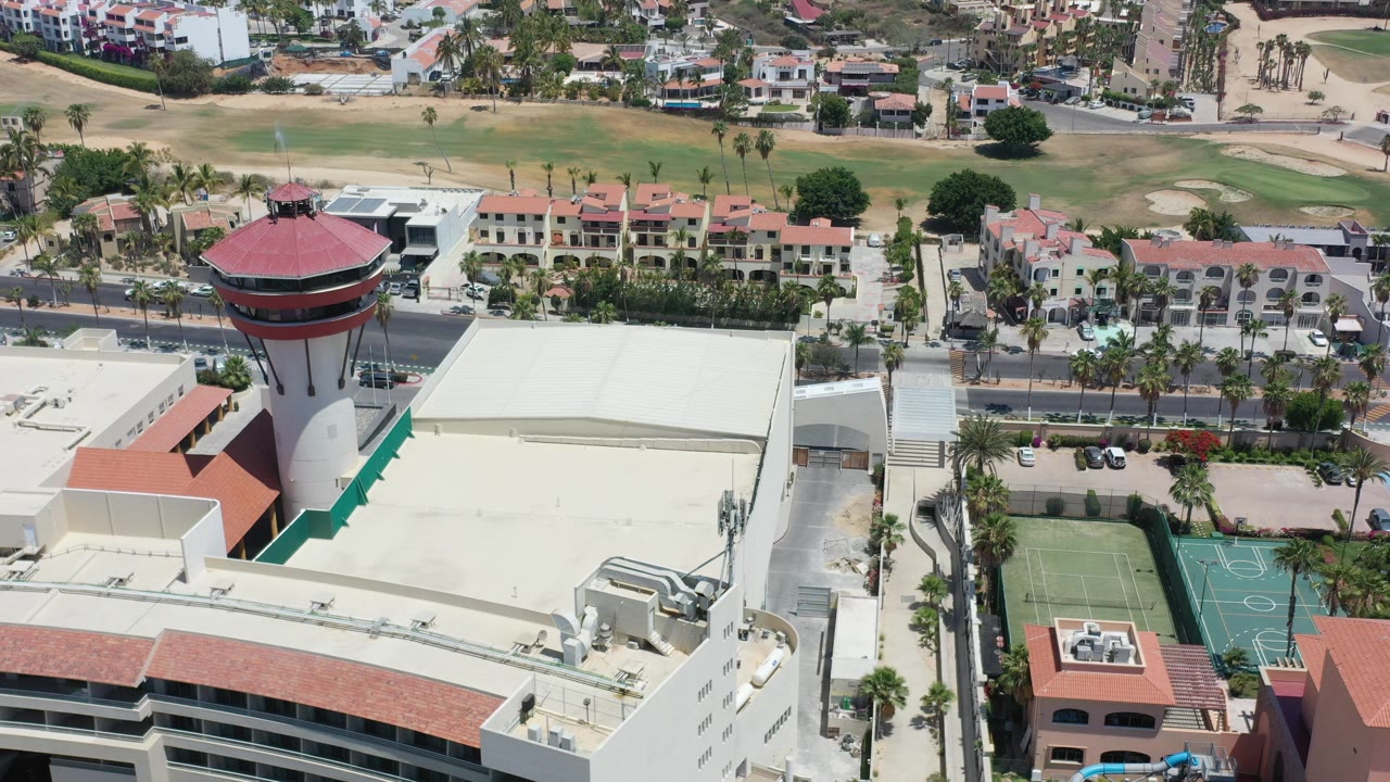High aerial frame over Los Cabos, Mexico showing a red-roofed round tower at left, pale rooftops, sport courts, a road, villas, palm trees, and golf fairways, with no visible people.