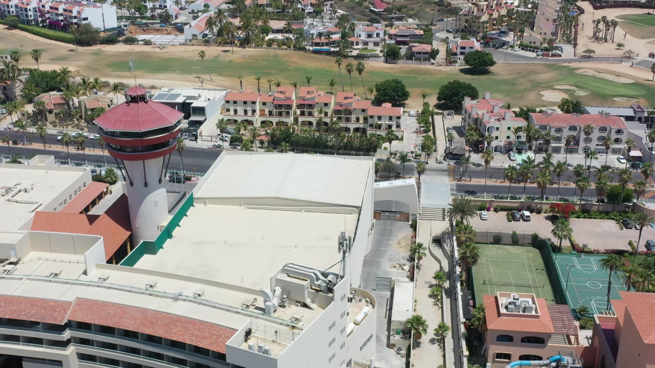 Aerial Los Cabos view with the red tower nearer the center-left, a large pale roof in the foreground, green sport courts, a palm-lined road, low villas, and golf fairways beyond, with no visible people.