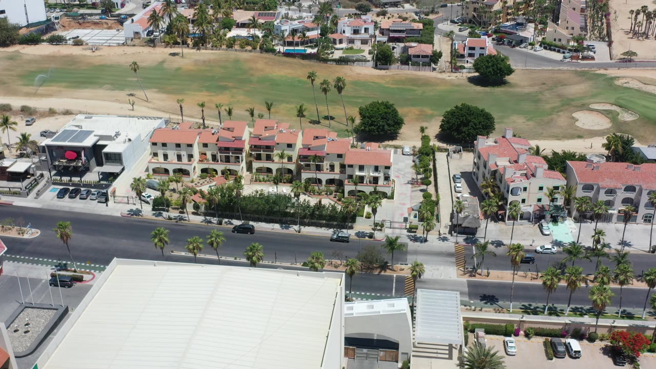 Aerial view in Los Cabos, Mexico, showing a few cars on the road, palms along the median and sidewalk, low-rise resort buildings, a black storefront sign area, and no visible people.