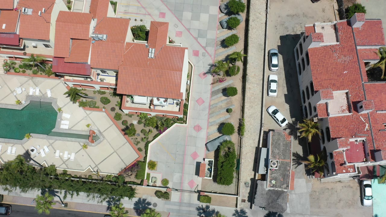 Aerial frame over Los Cabos residential buildings with red roofs, a green pool, white lounge chairs, sidewalk patterns, parked cars, palms, and no visible people.
