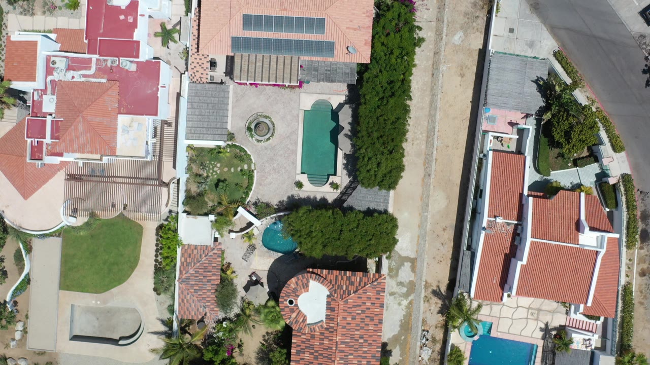 Overhead view of homes, lawns, trees, pools, and a road.
