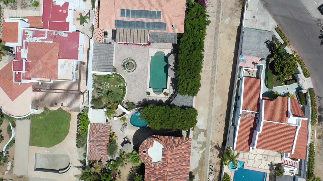 Overhead Los Cabos view centered on tiled-roof houses, a curved street edge, small pools, lawns, and dense green trees, with no visible people.