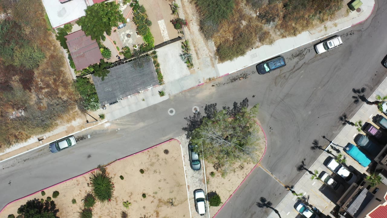 Overhead Los Cabos, Mexico view with a broad curve in the road, a red-roofed home at the edge, parked vehicles, sparse vegetation, and no visible people.
