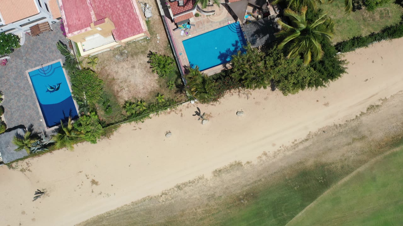 Second overhead frame in Los Cabos, Mexico with the shoreline lower in frame, red-tile roofs, bright blue pools, palms, beach sand, and no visible people.
