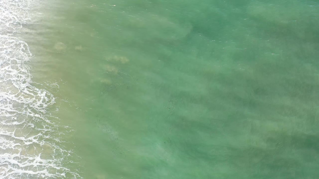 Aerial view over Los Cabos coastal water, where a thin white surf band runs down the left side and no visible people appear.