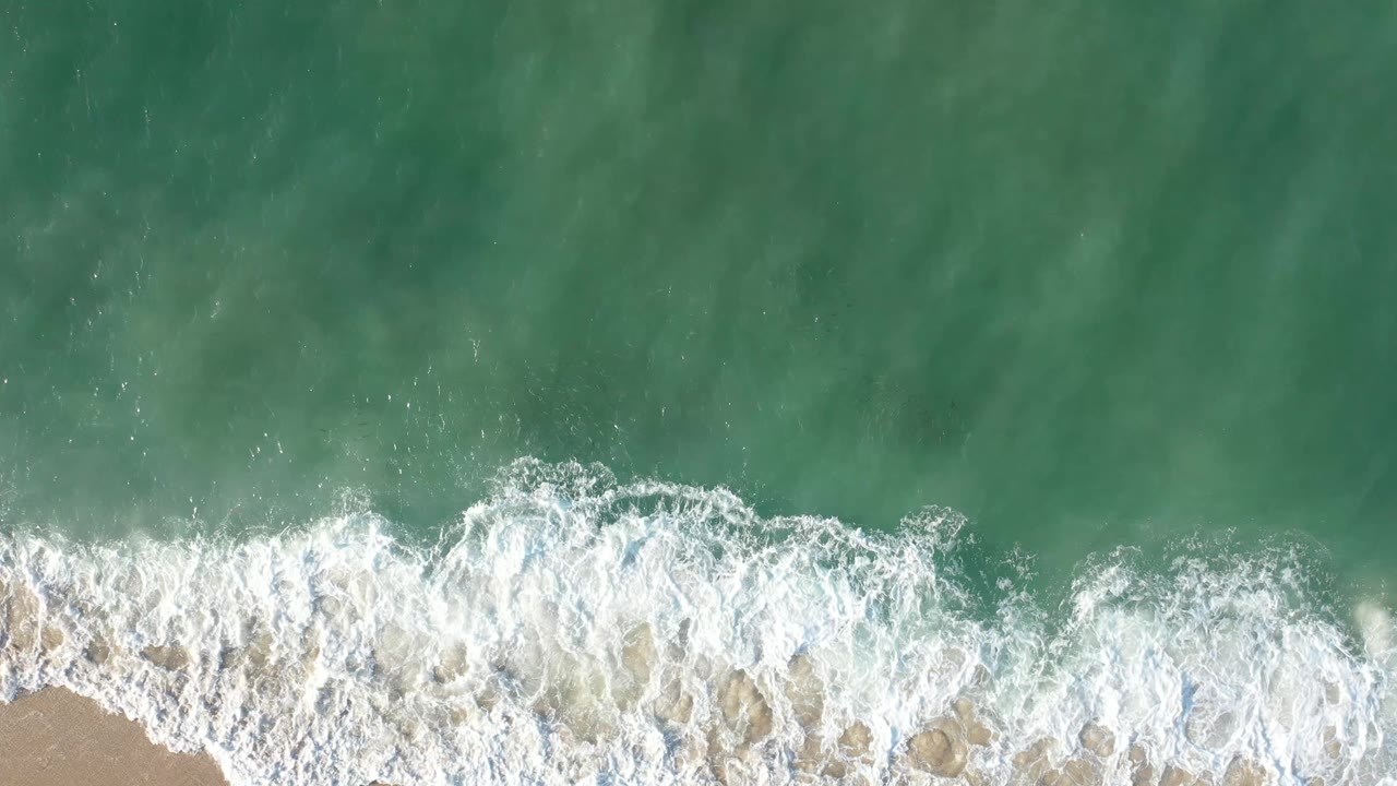 Aerial frame of white foam curling across green water near the sandy shoreline in Los Cabos, Mexico, with no visible people.