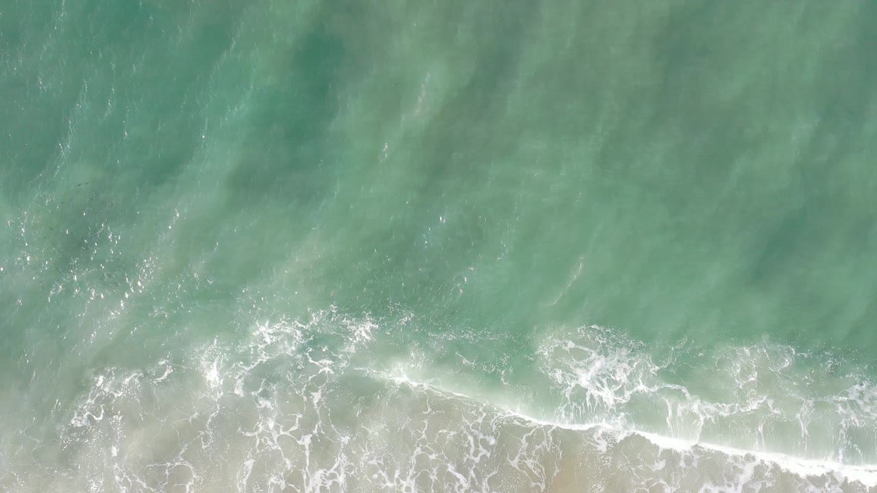Overhead view in Los Cabos, Mexico of green turquoise water and thin white foam reaching the sandy beach, with no visible people.