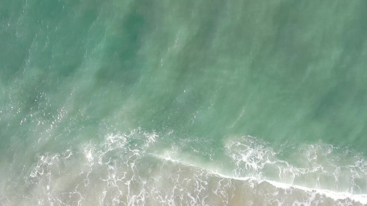 Aerial Los Cabos shoreline frame with a brighter band of white foam spreading over pale sand below green water, with no visible people.